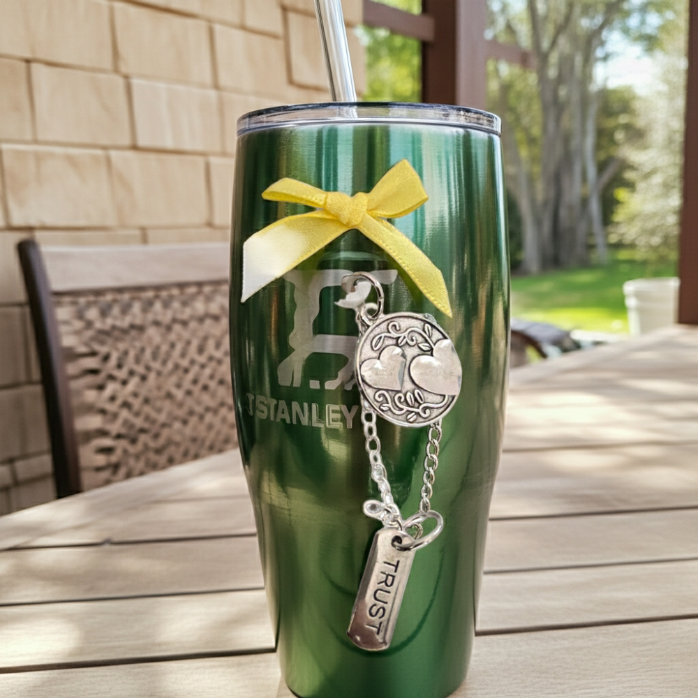 Green Stanley tumbler with a yellow ribbon and charm on a wooden table outdoors.